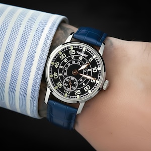 May include: A silver wristwatch with a black face and blue leather band. The watch face displays white numerals and hands, with a smaller subdial. The watch is on a wrist, with a blue and white striped shirt sleeve visible.