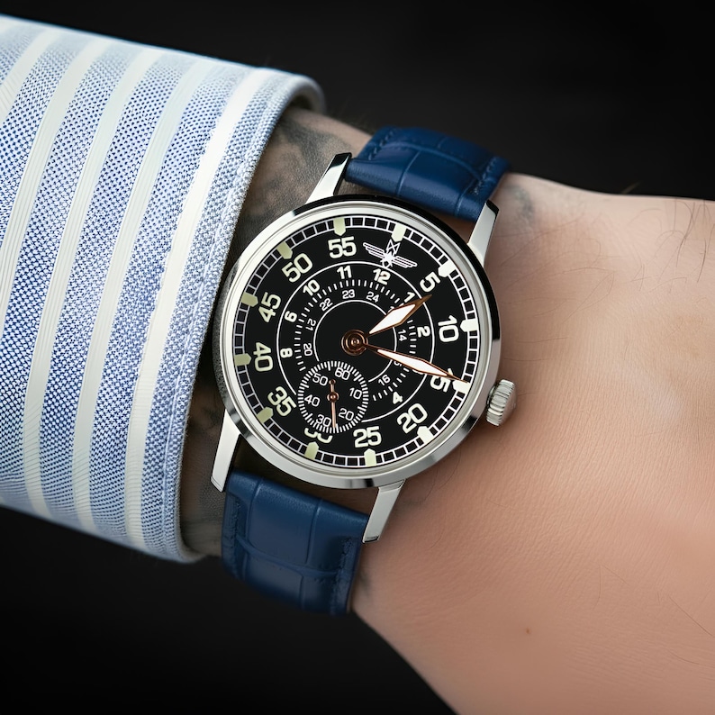 May include: A silver wristwatch with a black face and blue leather band. The watch face displays white numerals and hands, with a smaller subdial. The watch is on a wrist, with a blue and white striped shirt sleeve visible.