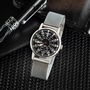 May include: A silver wristwatch with a black face and a mesh band. The watch face displays white numerals and hands, with a smaller dial at the bottom. A black pen is visible in the background.