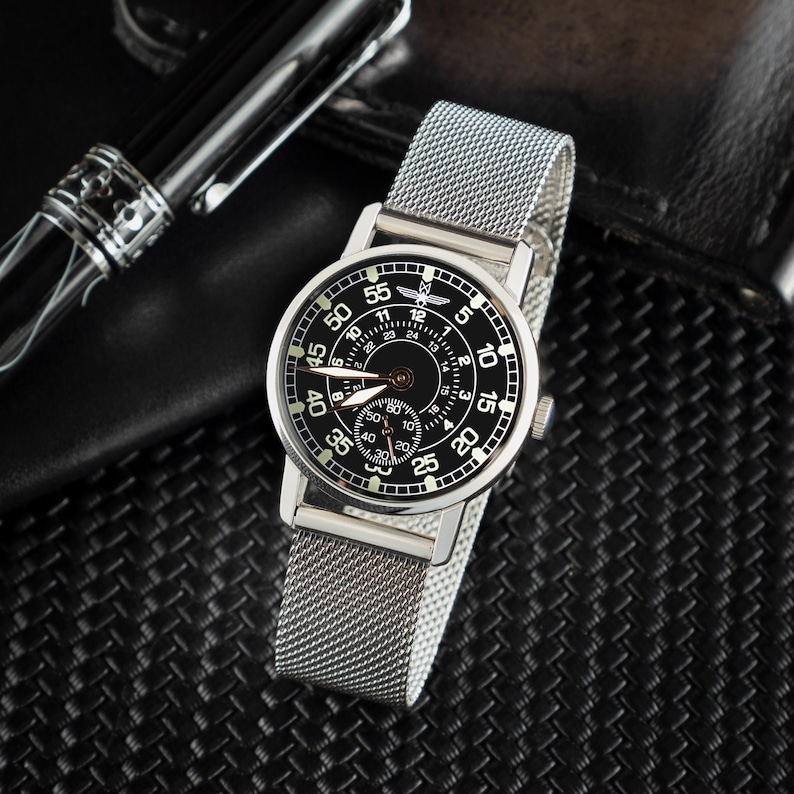 May include: A silver wristwatch with a black face and a mesh band. The watch face displays white numerals and hands, with a smaller dial at the bottom. A black pen is visible in the background.