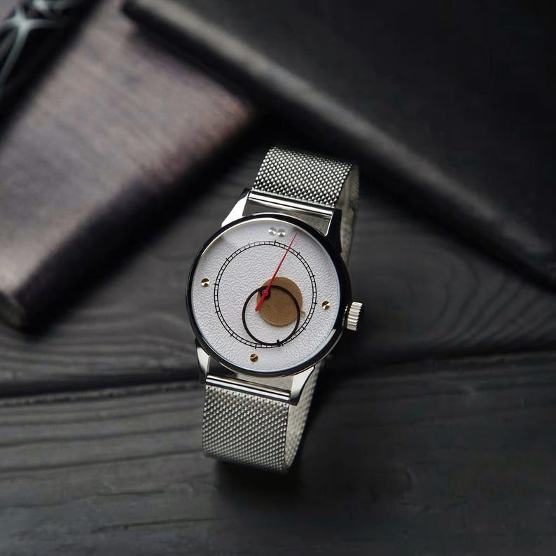 K&ouml;nnte beinhalten: Eine silberne Armbanduhr mit einem Mesh-Armband und einem wei&szlig;en Zifferblatt. Das Zifferblatt hat einen schwarzen Ring, einen braunen Kreis und einen roten Zeiger. Die Uhr liegt auf einer dunklen Holzoberfl&auml;che.