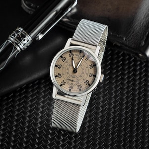 May include: A silver wristwatch with a mesh band. The watch face is beige with black numerals and hands. The watch is set against a dark background, with a pen visible in the upper left corner.