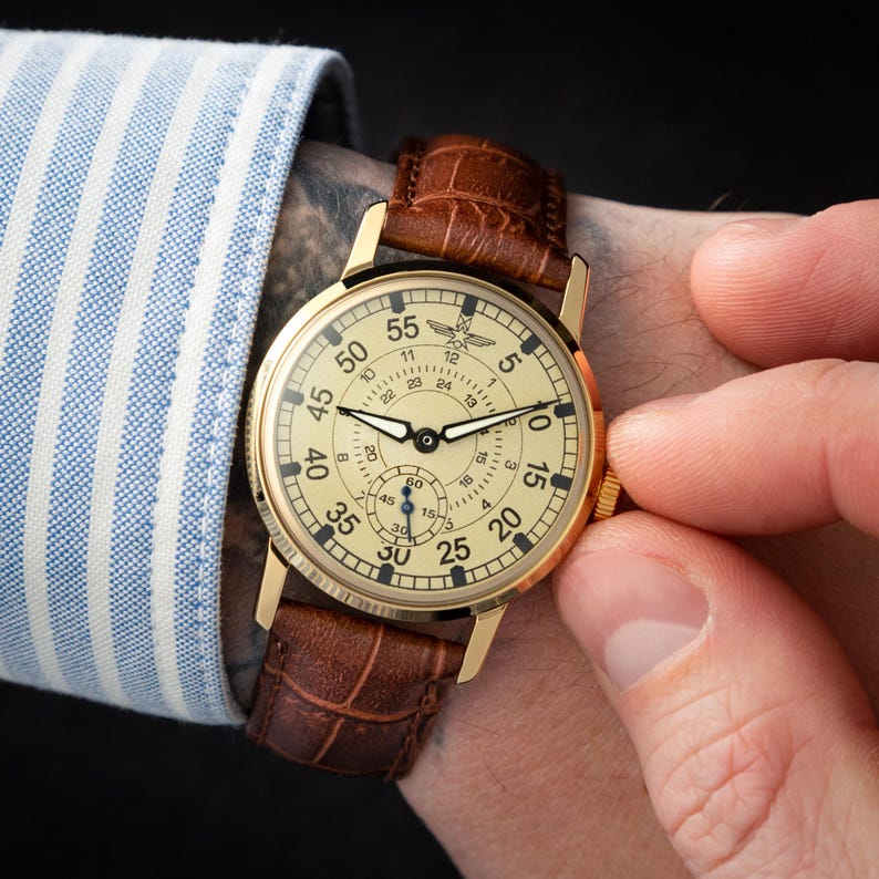 May include: A gold-toned wristwatch with a cream-colored face and brown leather band. The watch face displays black numerals and a smaller subdial. The watch is being adjusted by a person wearing a blue and white striped shirt.