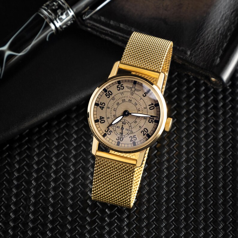 May include: A gold-toned wristwatch with a mesh band. The watch face has black numerals and hands, with a smaller subdial. The watch is set against a textured black surface, with a pen and wallet in the background.