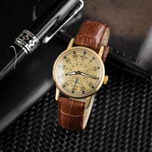 May include: A gold-toned wristwatch with a cream-colored face and brown leather band. The watch face displays black numerals and hands, with a smaller dial at the bottom. The band has a textured, crocodile-like pattern.