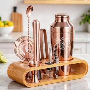 Op de afbeelding: Een rosé gouden cocktailset met een shaker, zeef, jigger, muddler, tang, flesopener en lepel, gepresenteerd in een bamboehouder. De shaker is gegraveerd met de naam "SPENCER".