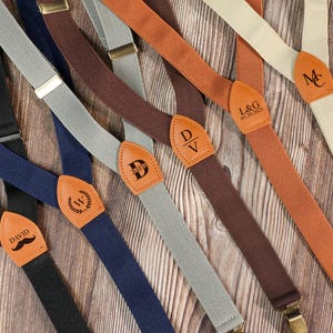 Puede incluir: Tirantes variados en varios colores, incluyendo negro, azul marino, gris, marrón y beige. Cada tirante presenta un parche de cuero con iniciales o nombres personalizados. Los tirantes tienen cierres metálicos y están sobre una superficie de madera.