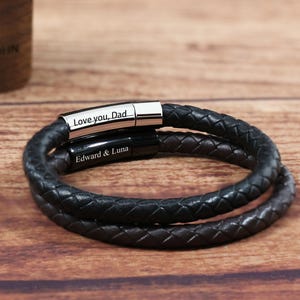 Puede incluir: Una pulsera de cuero negro con cierre plateado. La pulsera está grabada con las palabras "Love you, Dad" y "Edward & Luna". La pulsera se muestra sobre una superficie de madera.