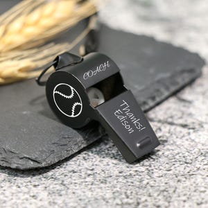 Könnte beinhalten: Eine schwarze Baseball-Metallpfeife mit dem Wort "COACH" und einer Baseball-Grafik. Die Pfeife trägt den Text "Thanks! Edison" auf der Seite. Die Pfeife liegt auf einer dunkelgrauen Oberfläche mit einem Weizenhalm im Hintergrund.