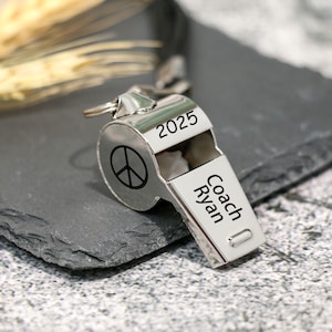 Könnte beinhalten: Eine silberne Trillerpfeife mit dem eingravierten Text "2025" und "Coach Ryan". Die Trillerpfeife hat ein Friedenssymbol. Die Trillerpfeife liegt auf einer dunkelgrauen Oberfläche.