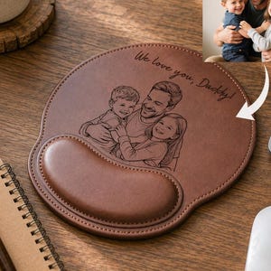 May include: A round brown leather mouse pad with a wrist rest, featuring a laser-engraved line drawing of a parent and two children hugging, with the text "We love you, Daddy!" inscribed above the illustration on the surface of the pad.