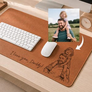 May include: A rectangular tan leather desk mat featuring a laser-engraved illustration of a person carrying a child on their shoulders. The text "I love you, Daddy!" is engraved in a cursive script next to the illustration on the mat surface.
