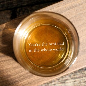 May include: A round glass filled with a golden liquid, set on a wooden surface. The text "You're the best dad in the whole world" is displayed inside the glass. The image is captured from above, showing the glass and its contents.