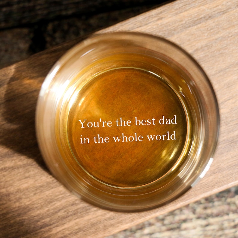 May include: A round glass filled with a golden liquid, set on a wooden surface. The text "You're the best dad in the whole world" is displayed inside the glass. The image is captured from above, showing the glass and its contents.