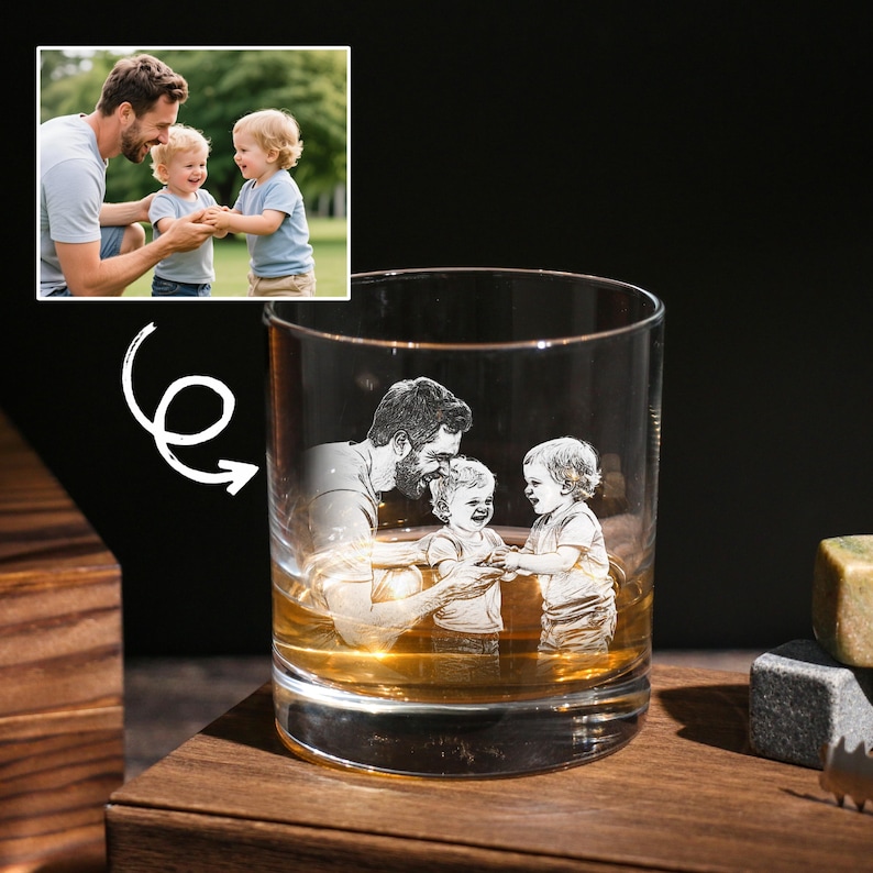 May include: A clear rocks glass featuring an engraved image of a father and two children. The glass contains amber liquid and rests on a wooden surface. A thoughtful gift idea.