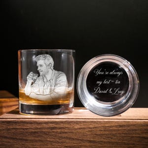 May include: A clear glass tumbler featuring an etched portrait of a man with a drink. Next to it, a clear glass lid with the inscription "You're always my best - tea David & Lucy". The glass is on a wooden surface.