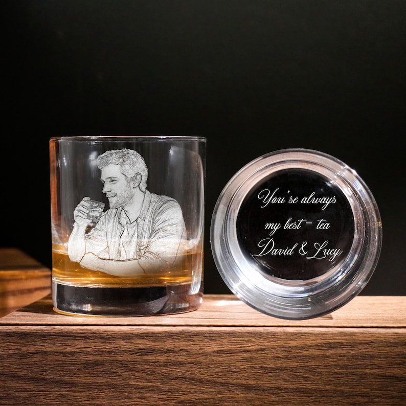 May include: A clear glass tumbler featuring an etched portrait of a man with a drink. Next to it, a clear glass lid with the inscription "You're always my best - tea David & Lucy". The glass is on a wooden surface.