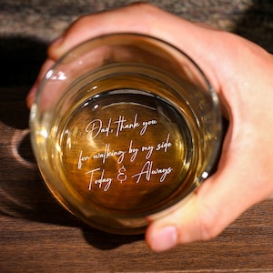 May include: A clear whiskey glass filled with amber liquid, held in a hand. The glass features an engraved message: "Dad, thank you for walking by my side Today & Always". The glass sits on a wooden surface.