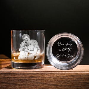 May include: A clear whiskey tumbler with an etched image of a person and a dog, filled with amber liquid. Next to it, a clear coaster with the text "You're always my best Tea David & Lucy" in white script. The items are on a wooden surface.