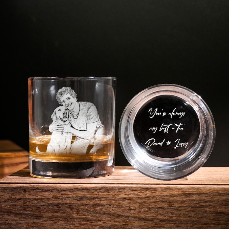 May include: A clear whiskey tumbler with an etched image of a person and a dog, filled with amber liquid. Next to it, a clear coaster with the text "You're always my best Tea David & Lucy" in white script. The items are on a wooden surface.