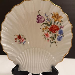 Op de afbeelding: Een witte, schelpvormige decoratieve schaal met een geschulpte rand en gouden rand. De schaal heeft een handgeschilderd bloemmotief in tinten rood, paars, geel en blauw. Het wordt weergegeven op een zwarte standaard.