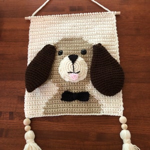Puede incluir: Un tapiz de pared de ganchillo con un perro marrón y blanco con una pajarita negra. El perro tiene orejas marrones, ojos negros y una lengua rosa. El tapiz tiene cuentas de madera y borlas.
