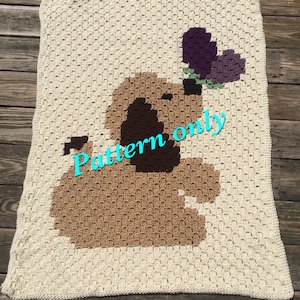 May include: A crochet pattern for a brown dog with a purple flower on its head. The pattern is worked in squares and is suitable for beginners. The text "Pattern only" is written on the image.