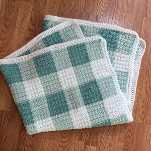 May include: A handmade crochet blanket with a checkered pattern in shades of green and white. The blanket has a textured appearance and a cream-colored border. The blanket is folded and resting on a wooden surface.