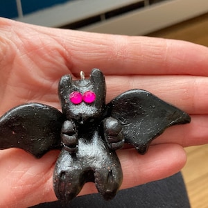 May include: A black bat charm with pink eyes. The bat is made of polymer clay and has a textured surface.