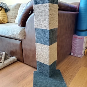 Puede incluir: Un rascador para gatos con base cuadrada y una columna alta y multicolor. La columna alterna secciones de material tejido gris y beige. La base es un cuadrado gris sólido. El poste está diseñado para que los gatos rasquen.