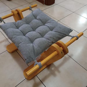 May include: A small, wooden pet bed with a gray, tufted cushion. The bed has a simple design with two wooden supports and a rectangular base. The cushion is secured with gray rope. The bed is designed for small pets.