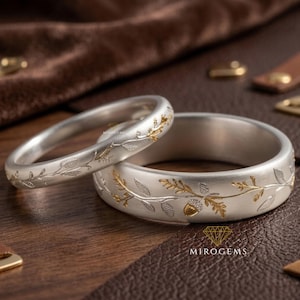 Op de afbeelding: Twee zilveren ringen met gouden blad- en eikelontwerpen. De ringen liggen op een bruin oppervlak. De ringen hebben een matte afwerking en zijn waarschijnlijk van metaal. De ringen zijn waarschijnlijk voor een bruiloft of verloving.