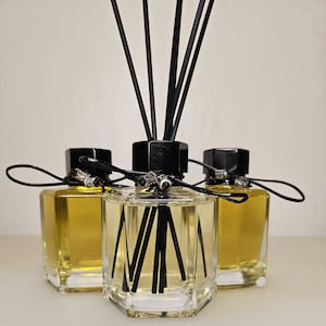 May include: Three hexagonal glass reed diffuser bottles filled with a golden liquid. Each bottle features a black cap, black reed sticks, and a black cord tied around the neck with a small silver-toned decorative bead accenting the cord.