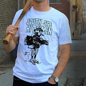 Puede incluir: Camiseta blanca con una ilustraci&oacute;n en blanco y negro de una persona que lleva una bolsa de golf y un palo. El texto "South Side Hitmen" est&aacute; impreso en la camiseta.