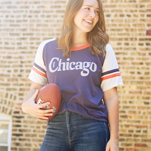 May include: A woman wearing a purple and white baseball jersey with the word "Chicago" on the front. She is holding a brown football in her left hand and is wearing blue jeans.
