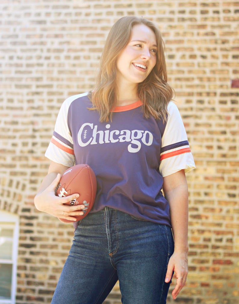 May include: A woman wearing a purple and white baseball jersey with the word "Chicago" on the front. She is holding a brown football in her left hand and is wearing blue jeans.