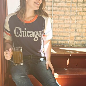 May include: A woman wearing a blue and white baseball jersey with the word "Chicago" on the front. She is sitting on wooden steps and holding a glass mug of a light brown drink.