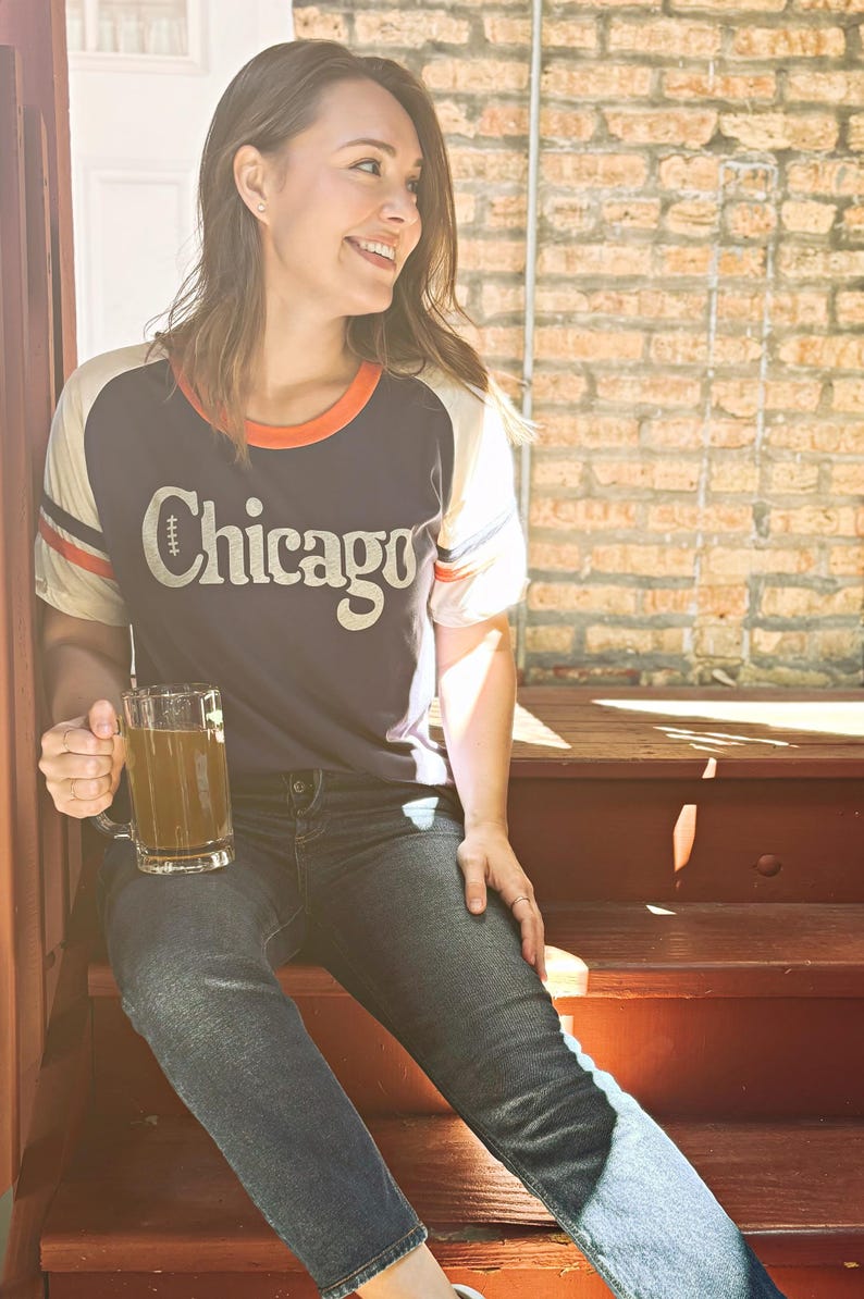 May include: A woman wearing a blue and white baseball jersey with the word "Chicago" on the front. She is sitting on wooden steps and holding a glass mug of a light brown drink.