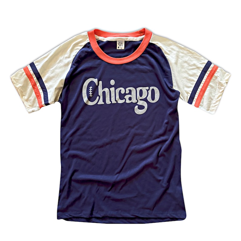 May include: A navy blue t-shirt with a white and red baseball-style striped sleeve. The shirt has a white "Chicago" graphic with a football graphic above the text.