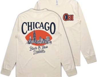 May include: A beige long-sleeve t-shirt with a graphic of the Chicago skyline and the text "CHICAGO" in a vintage font. The back of the shirt has the text "Black & Blue Division" and a football graphic. The front of the shirt has the text "CHI" in a vintage font.