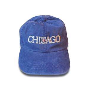 May include: A blue baseball cap with the word "CHICAGO" embroidered in white across the front. The "O" in "CHICAGO" features a baseball design. The cap has a curved brim and a button on top.