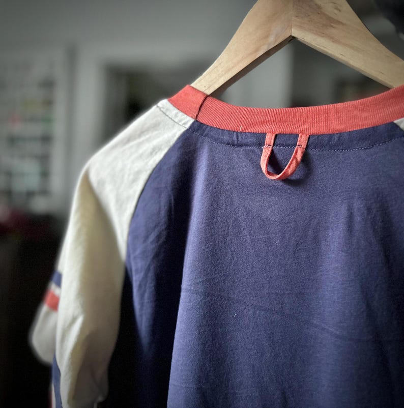 May include: A navy blue and white baseball tee shirt with a red trim around the neck. The shirt has a loop on the back of the neck.