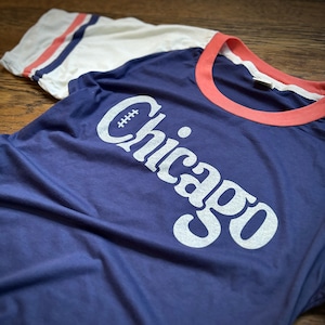 May include: A navy blue t-shirt with a white ringer and white sleeves. The shirt has a white graphic of the word "Chicago" with a football in the "C".