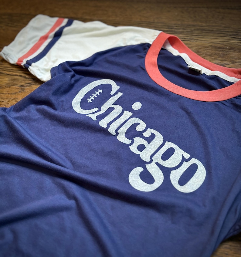 May include: A navy blue t-shirt with a white ringer and white sleeves. The shirt has a white graphic of the word "Chicago" with a football in the "C".