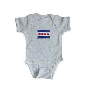 May include: A light gray baby onesie with a Chicago flag patch. The patch features a blue stripe at the top and bottom, with three red six-pointed stars in the center. The onesie has short sleeves and a snap closure.