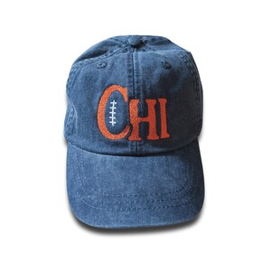 Peut inclure: Casquette de baseball bleue avec le mot "CHI" brodé en orange. Le "C" est en forme de ballon de football américain. La casquette a une visière incurvée et un bouton sur le dessus.