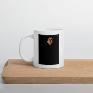 Può includere: Una tazza in ceramica bianca con una stampa grafica rettangolare sul davanti. La stampa mostra il ritratto di una persona dai capelli scuri con un'espressione seria, su uno sfondo nero profondo che occupa la maggior parte dell'immagine.