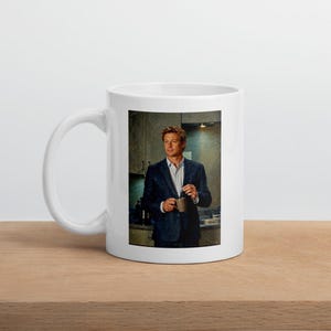 Può includere: Una tazza in ceramica bianca con l'immagine stampata di una persona con capelli biondi corti che indossa un blazer blu scuro e una camicia azzurra. La persona tiene in mano una piccola tazza marrone in una cucina con armadietti scuri.