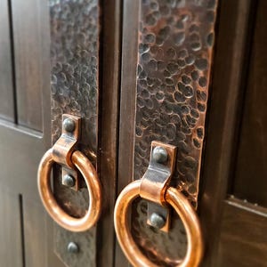May include: Two hammered copper door handles with ring pulls. The handles are mounted on a dark brown wooden door.