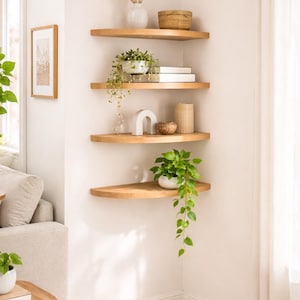 Könnte beinhalten: Vier Eckregale aus hellem Holz, die an einer weißen Wand montiert sind. Die Regale tragen verschiedene Dekorationsgegenstände, darunter eine weiße Vase mit Trockenblumen, einen geflochtenen Korb, grüne Topfpflanzen, Bücher und ein weißes Bogenobjekt.